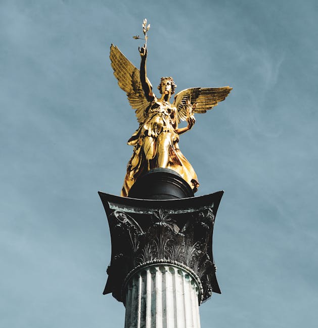 A close-up view of a tall, classical column with a detailed capital, supporting a golden angel sculpture with outstretched wings and a raised arm holding an object, set against a clear blue sky. The sculpture appears to be made of gilded material, contrasting with the dark base and the white marble of the column. This image captures the intricate design and grandeur of historic monument decoration, resembling elements typical of municipal or commemorative statues often seen in central London. The composition highlights the verticality and ornate craftsmanship typical of traditional European statue installations, relevant to service providers like Removal Company Islington involved in house removals and furniture transport, with the sculpture possibly located at a prominent public space or landmark in the Angel N1 area.