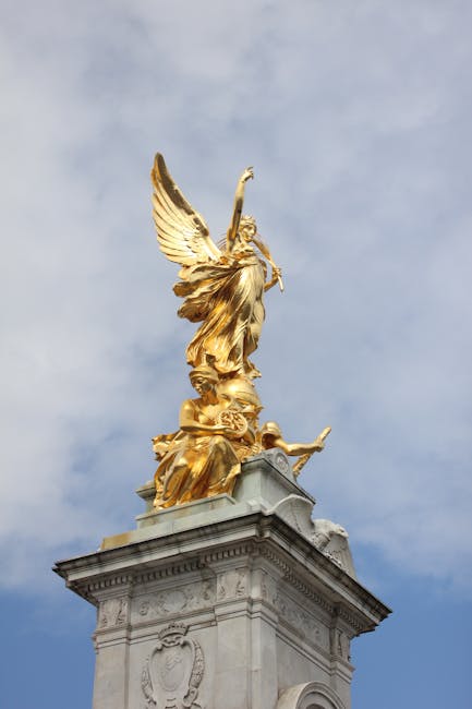 A detailed view of a tall, ornate pedestal topped with a gilded sculpture featuring a winged figure holding a staff, set against a partly cloudy sky. The sculpture is golden, with intricate detailing on the wings, clothing, and base, and is mounted on a stone base with decorative carvings. The scene appears to be outdoors, possibly at a monument or historical site, with the focus on the artistic and architectural elements. Although not related to house removals or moving services, this image captures the craftsmanship of a prominent gilded statue in a public space.