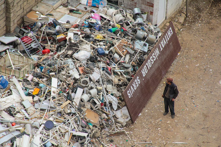 A person wearing a white shirt and beige trousers is holding a clear plastic bottle in their left hand, preparing for waste disposal or recycling as part of a home relocation process. Behind them, the
